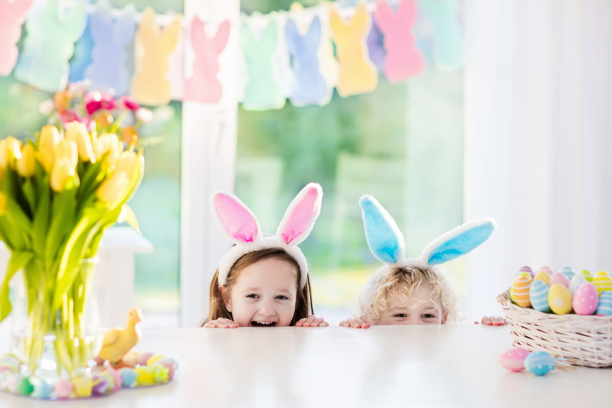 two kids wearing bunny ears peering over a kitchen counter at easter eggs