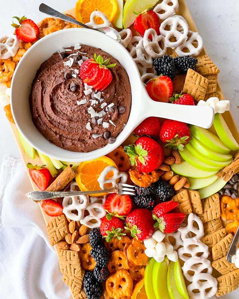 large wooden serving board with healthy fruit snacks and chocolate treats with a chocolate hummus dip