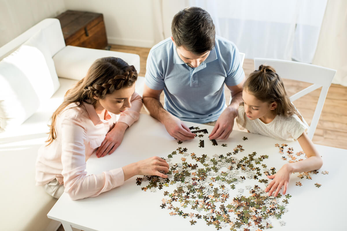 mother, father and daughter doing a puzzle on a white table in their living room