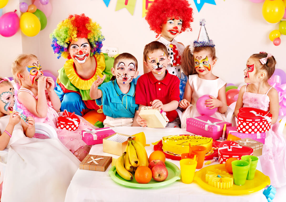 5 kids at a birthday party with two clowns and a table of food including a tray of fresh fruit